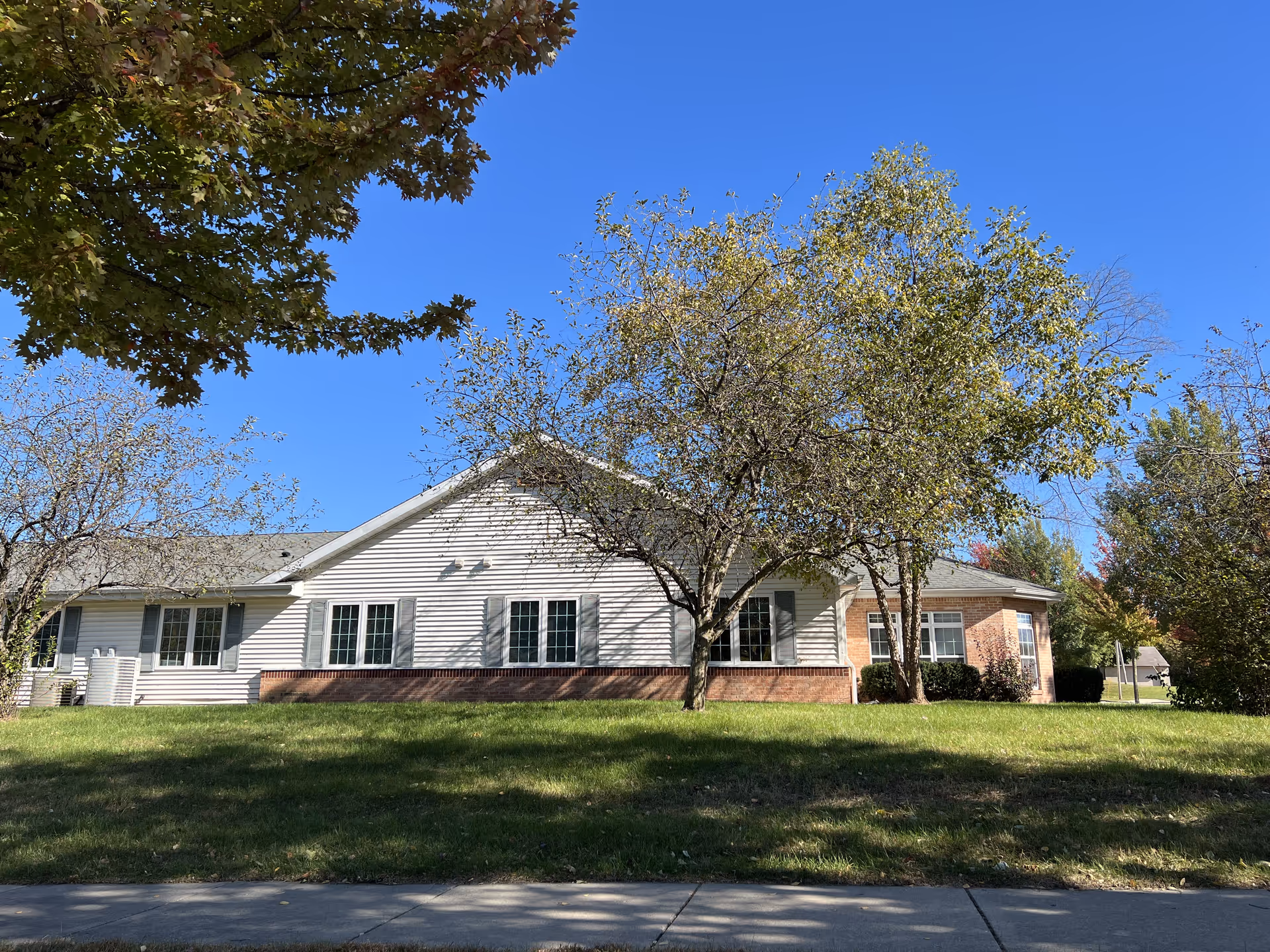 Exterior view of a single-story building with white siding and brick accents, surrounded by trees and green grass under a clear blue sky.