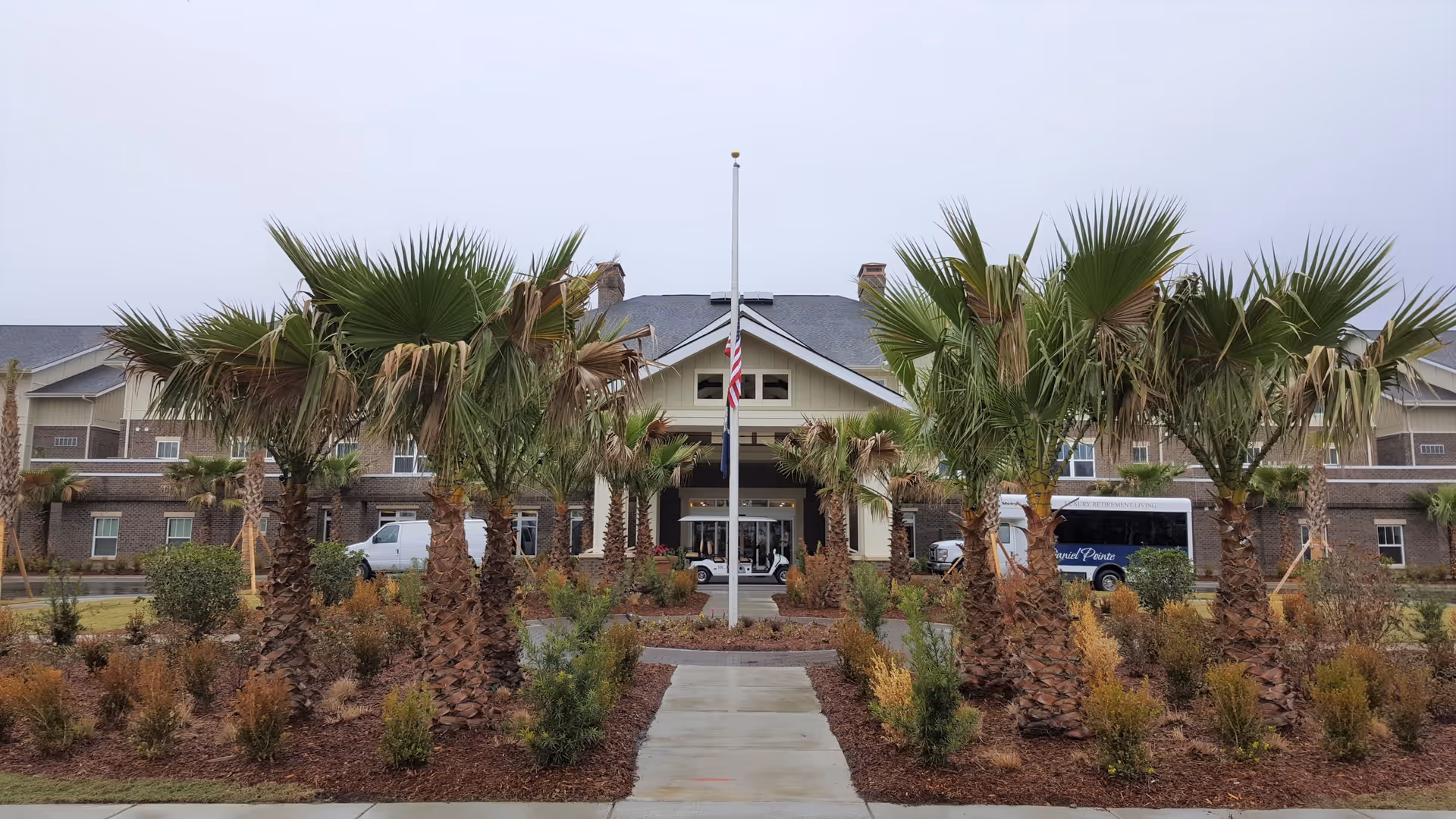 Front entrance of a retirement community building with a central flagpole, palm trees lining a walkway and a drive-up portico.