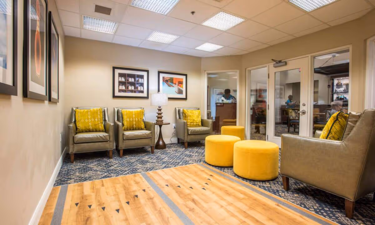 A cozy lounge area with four olive green armchairs each with a yellow patterned cushion, two round yellow ottomans, a wooden side table with a lamp, and framed artwork on the beige walls. The floor has a combination of patterned carpet and wood. Glass doors and windows reveal another room with people inside.