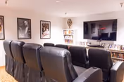 A cozy media room with several black leather recliner chairs arranged in rows facing a large flat-screen TV mounted on the wall. The room has framed movie posters on the walls and bookshelves filled with DVDs and books beneath the TV.