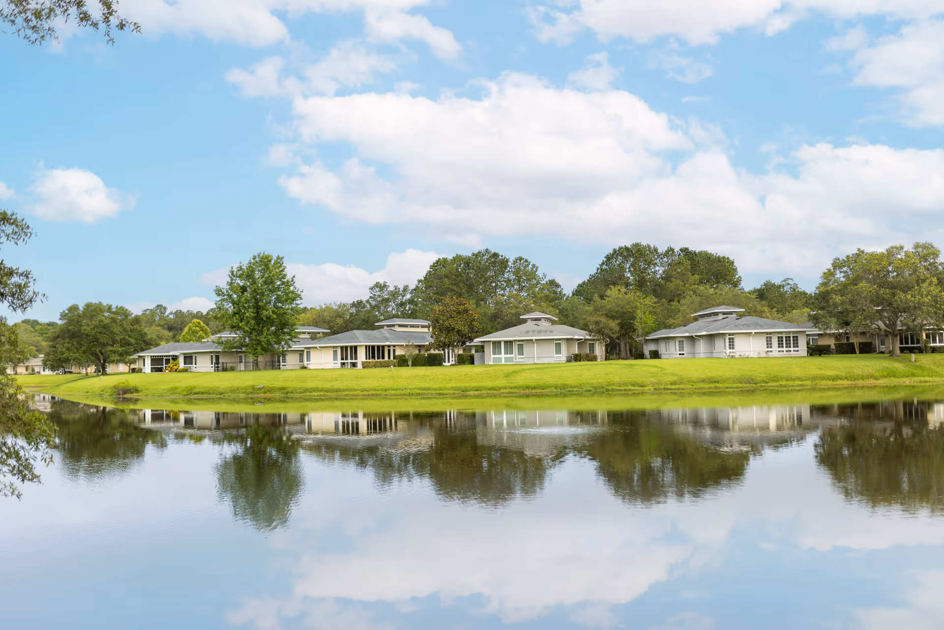 A serene view of several single-story buildings with light-colored walls and dark roofs situated along a grassy bank beside a calm body of water, reflecting the buildings, trees, and partly cloudy sky above.