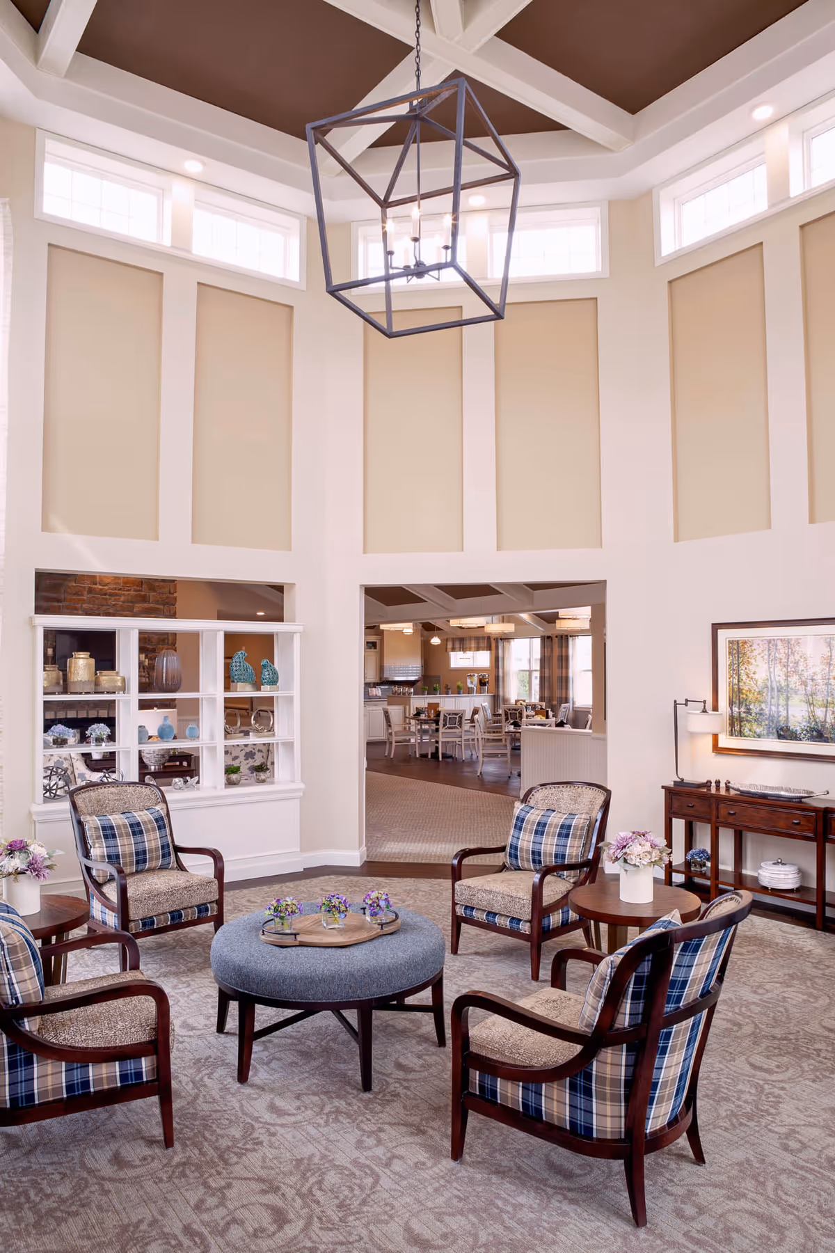 A cozy living room area with four wooden armchairs featuring plaid cushions arranged around a round upholstered ottoman with a wooden tray holding small flower arrangements. The room has high ceilings with a large geometric pendant light fixture and tall windows near the ceiling. In the background, there is a dining area with tables and chairs, and a decorative shelving unit with vases and ornaments.