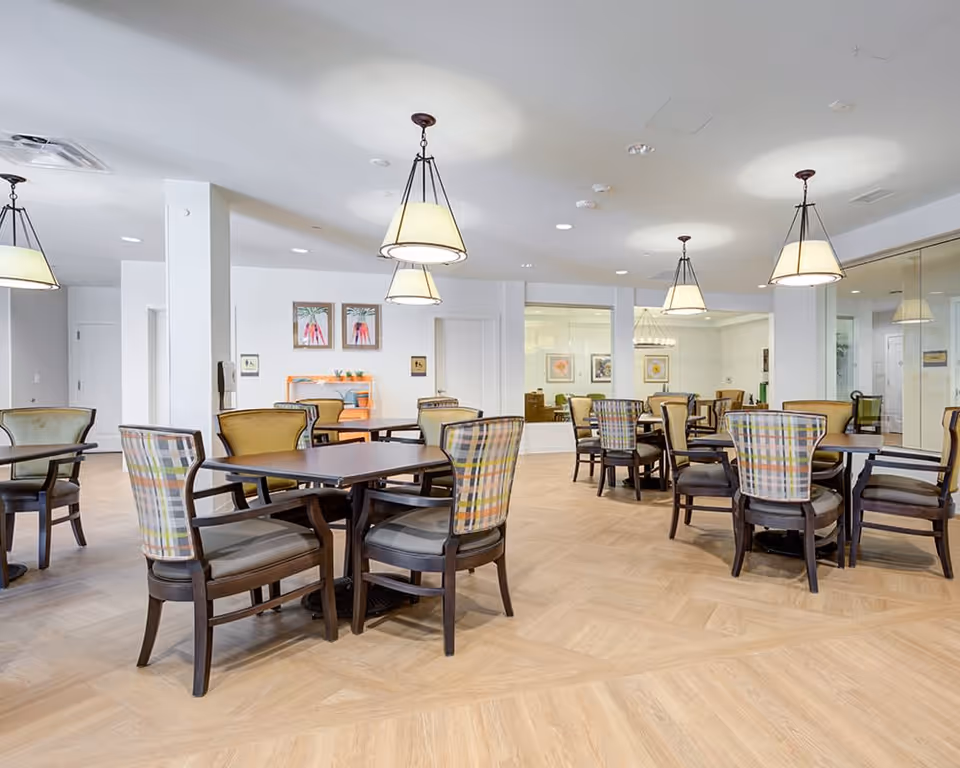 A bright and spacious dining room with multiple dark wood tables and chairs featuring colorful plaid upholstery. The room has light wood flooring, white walls, and several hanging pendant lights. There are framed artworks on the walls and a glass partition separating another seating area in the background.