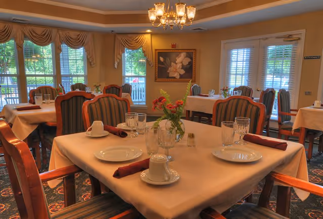 A warmly lit dining room with tables covered in beige tablecloths, each set with plates, glasses, cups, and red cloth napkins. The chairs have wooden frames with striped upholstery. There are windows with blinds and draped curtains letting in natural light, and a floral painting hangs on the wall.
