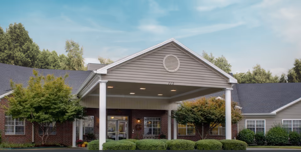 Front exterior view of Lincoln Manor facility showing a covered entrance with white columns, brick and siding walls, surrounded by green bushes and trees under a blue sky.