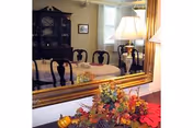 Dining room with a table and chairs reflected in a large mirror, a china cabinet, a lamp, and a floral centerpiece with small decorative pumpkins.