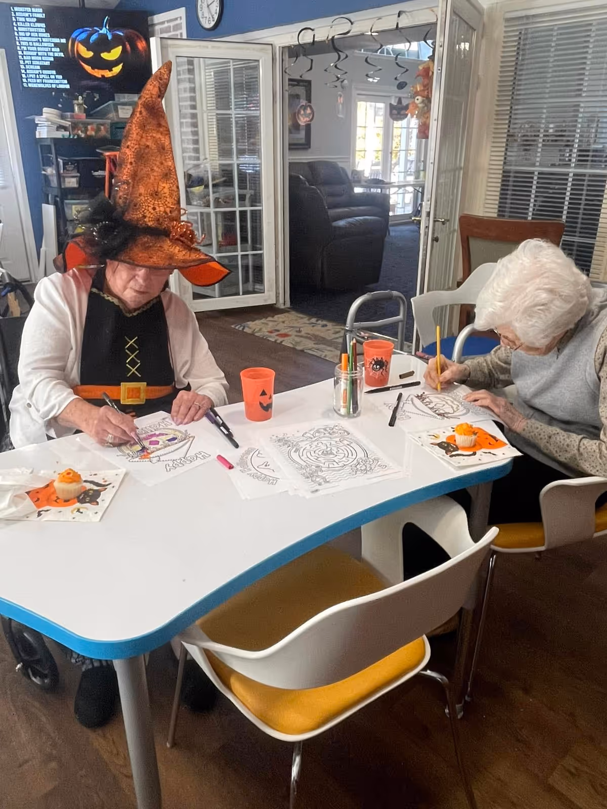 Two elderly women sitting at a white table engaged in Halloween-themed coloring activities. One woman is wearing an orange witch hat and a black outfit with a belt design, while the other is focused on coloring with a pencil. The table has coloring sheets, markers, orange cups with Halloween designs, and small Halloween-themed cupcakes on napkins. The room has a cozy interior with open French doors leading to a lounge area with chairs and Halloween decorations hanging from the ceiling.