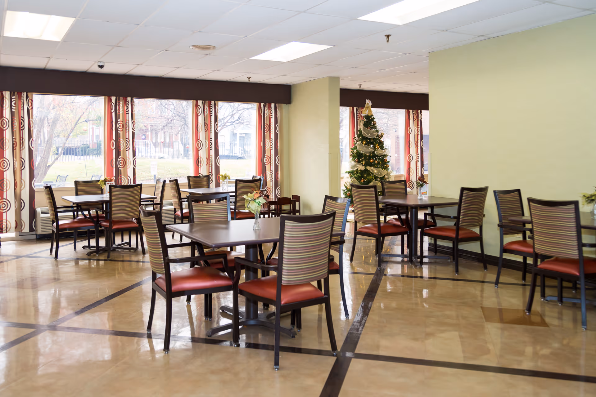 A bright dining room with multiple tables and chairs arranged neatly. The room has large windows with patterned curtains allowing natural light to enter. A decorated Christmas tree is visible near a corner, adding a festive touch to the space.
