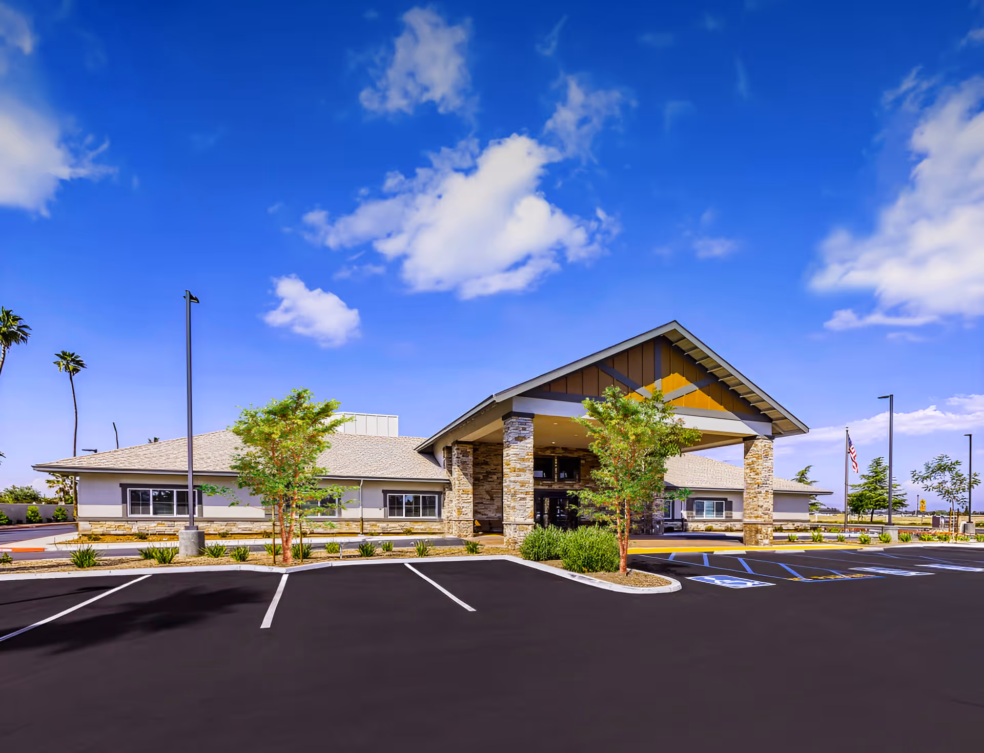 Exterior view of a single-story building with a covered entrance supported by stone pillars, surrounded by a parking lot with marked spaces and some small trees and shrubs. The sky is clear with a few clouds.