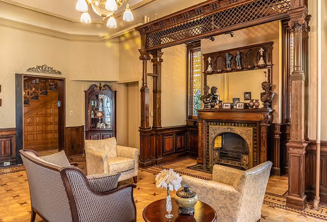A cozy living room with vintage wooden furniture and intricate woodwork. The room features a decorative fireplace with a large mirror above it, flanked by wooden columns and shelves holding small statues and framed photos. There are two upholstered armchairs and a wooden settee arranged around a small round table with a vase of flowers and decorative items. The room has warm lighting from a chandelier and a stained glass window beside the fireplace.