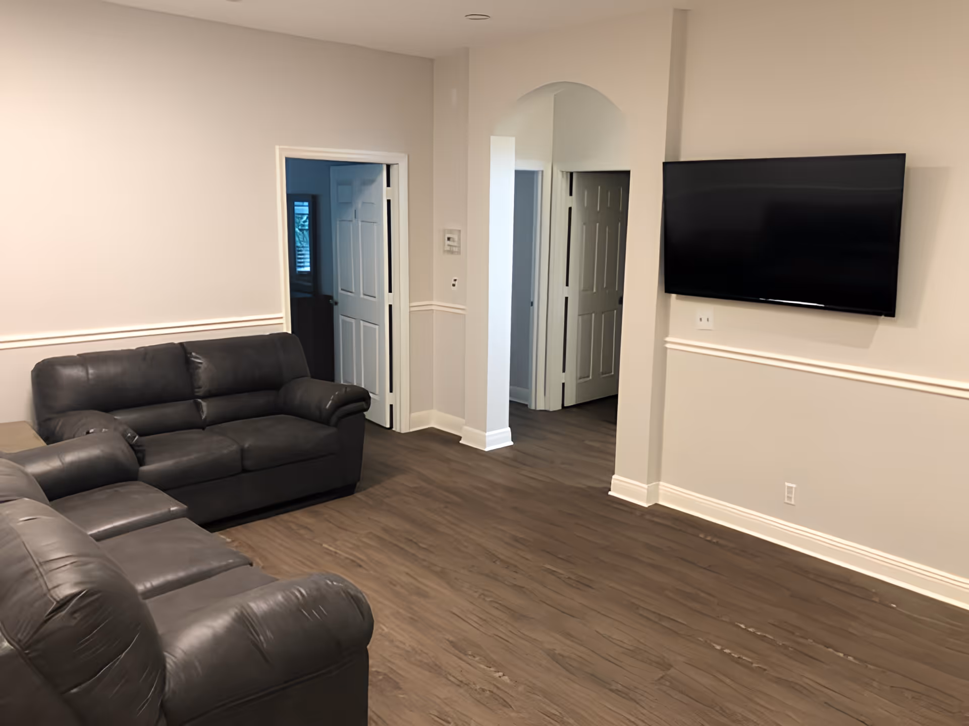 A living room with two dark gray leather sofas facing a wall-mounted flat screen TV. The room has light beige walls with white trim and dark wood flooring. There are two doorways and an arched opening leading to other rooms.