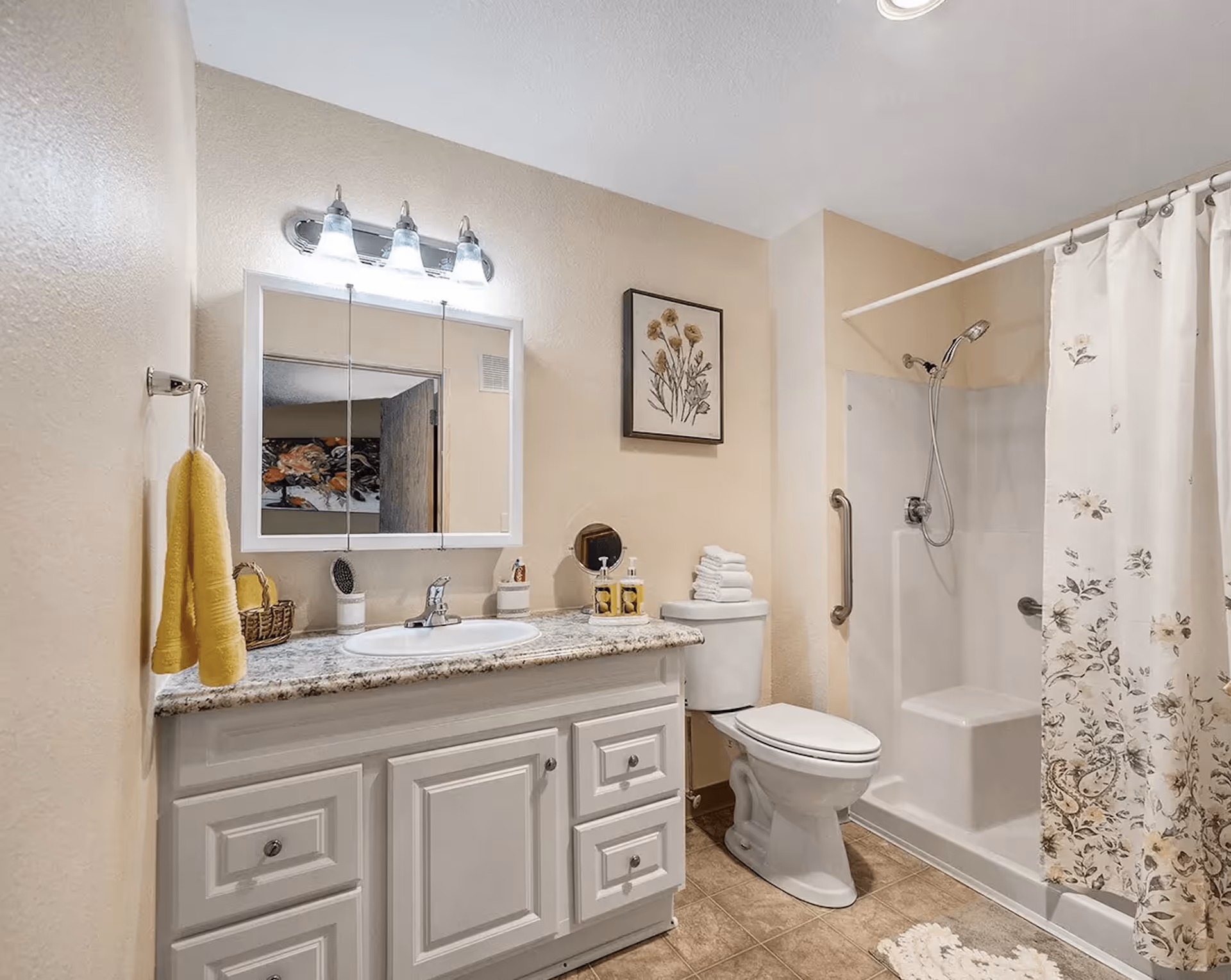 A clean and well-lit bathroom featuring a white vanity with granite countertop, a sink, a mirror with three lights above it, a toilet with folded towels on top, and a shower area with a floral-patterned curtain and a built-in seat.