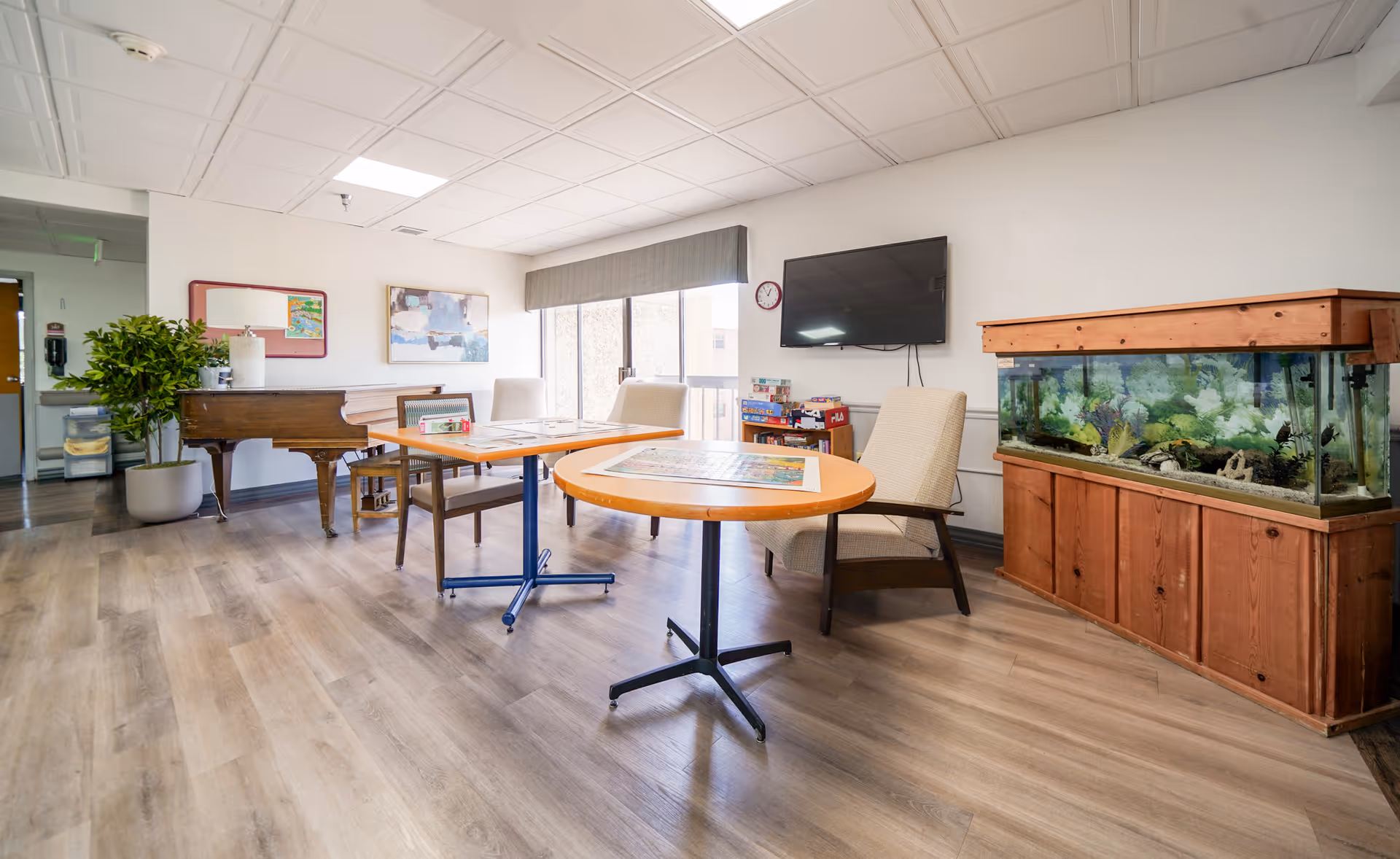 A bright and spacious common area in a senior living facility featuring two tables with chairs around them, a large fish tank on a wooden stand, a wall-mounted TV, a piano, a potted plant, and large windows letting in natural light.