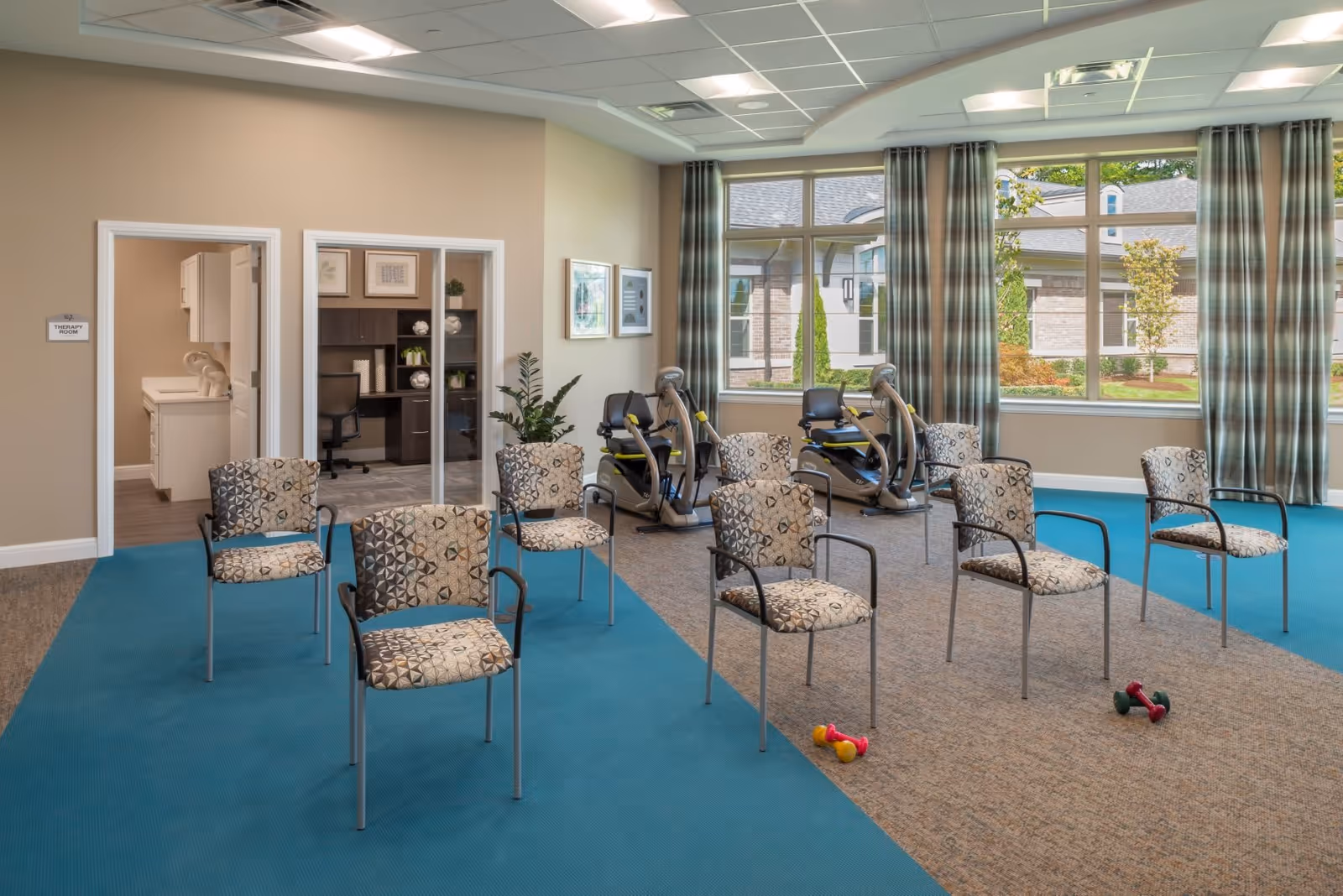 A therapy room in a senior living facility with several patterned chairs arranged in rows on a blue and beige carpet. There are two exercise bikes near large windows with plaid curtains, and small dumbbells on the floor. A door labeled 'Therapy Room' leads to an adjacent office area with a desk and shelving.