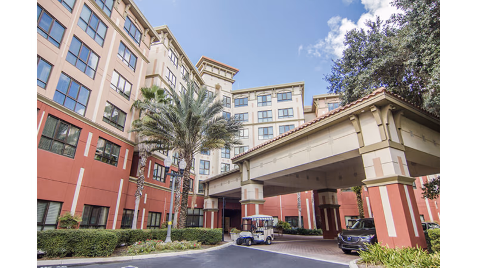 Exterior view of a multi-story senior living facility with a covered entrance, palm trees, and parked vehicles under a partly cloudy sky.