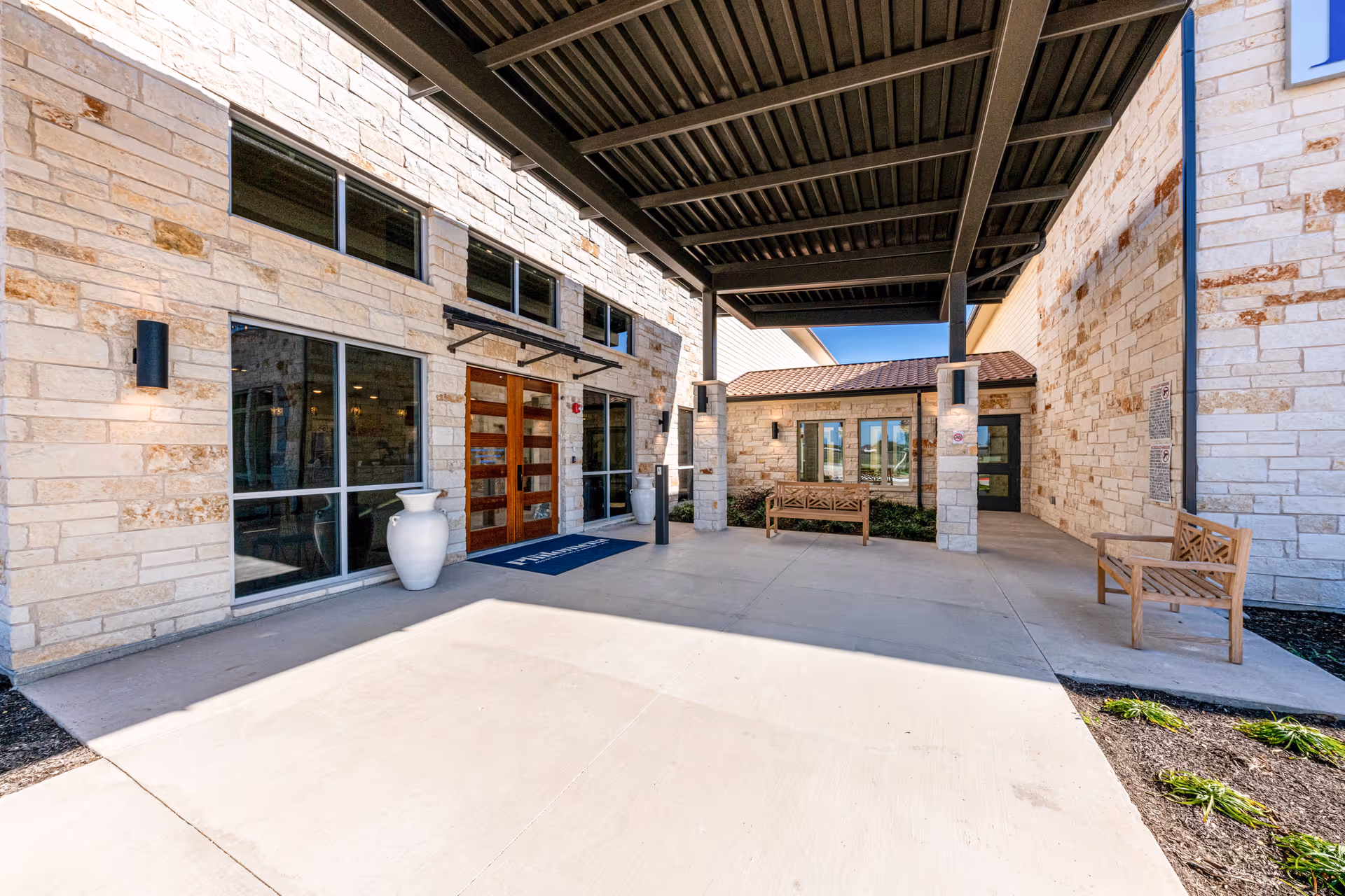 Covered entrance area of The Philomena facility with stone walls, large windows, wooden double doors, two large white decorative vases, and wooden benches on either side. The area is well-lit with wall-mounted lights and has a concrete floor with some landscaping visible on the right side.