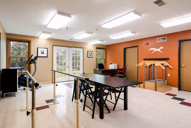 A rehabilitation or therapy room with parallel bars for walking exercises, a table with chairs, an exercise bike near a window, a desk with a chair and printer, and a small set of wooden stairs against an orange accent wall with a white running dog logo.