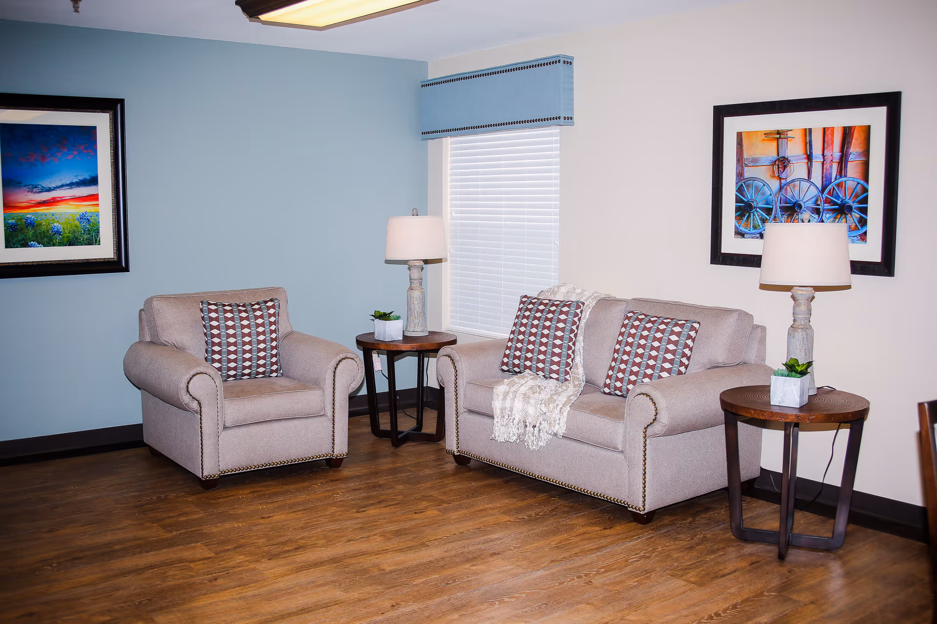 A cozy living area with an upholstered armchair and loveseat, side tables with lamps, patterned pillows, a throw, and framed artwork on the walls.