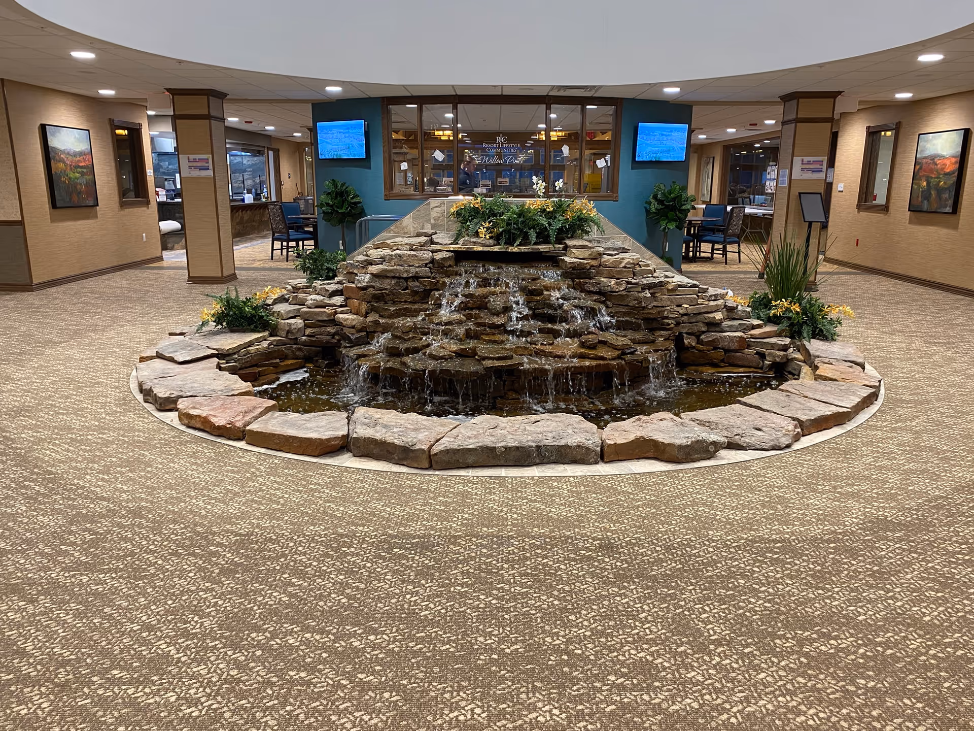 Carpeted retirement community lobby with a central stone water feature, plants, seating areas, and a reception window in the background.