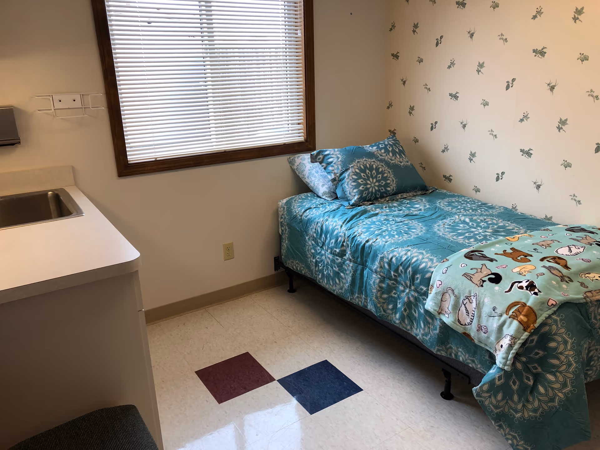 A small bedroom with a single bed covered in blue patterned bedding and a blanket with cat illustrations. The room has a window with closed blinds, a countertop with a sink, and wallpaper with a subtle floral pattern.