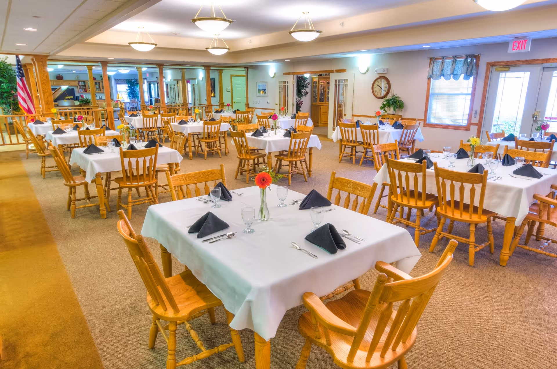 A bright dining room with multiple wooden tables and chairs arranged neatly. Each table is covered with a white tablecloth and set with black folded napkins, glasses, and silverware. There are small flower vases with colorful flowers on each table. The room has beige carpeting, light-colored walls, ceiling lights, and windows with curtains. An American flag is visible in the background near a wooden railing.