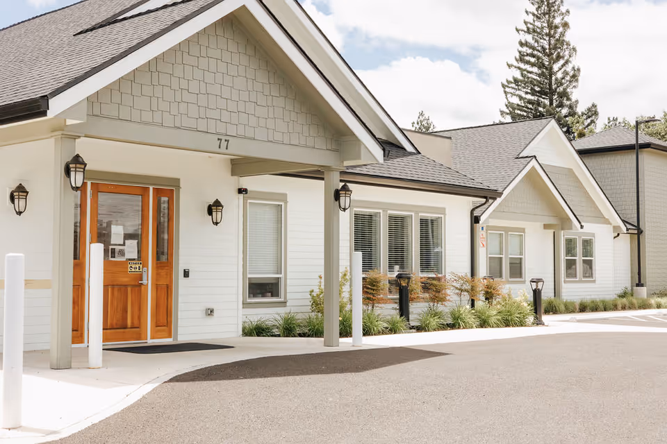 Exterior view of a single-story building with white siding and a gray shingled roof. The entrance features wooden double doors under a covered porch with the number 77 above. There are several windows with white blinds and small landscaped bushes along the front. The sky is partly cloudy.
