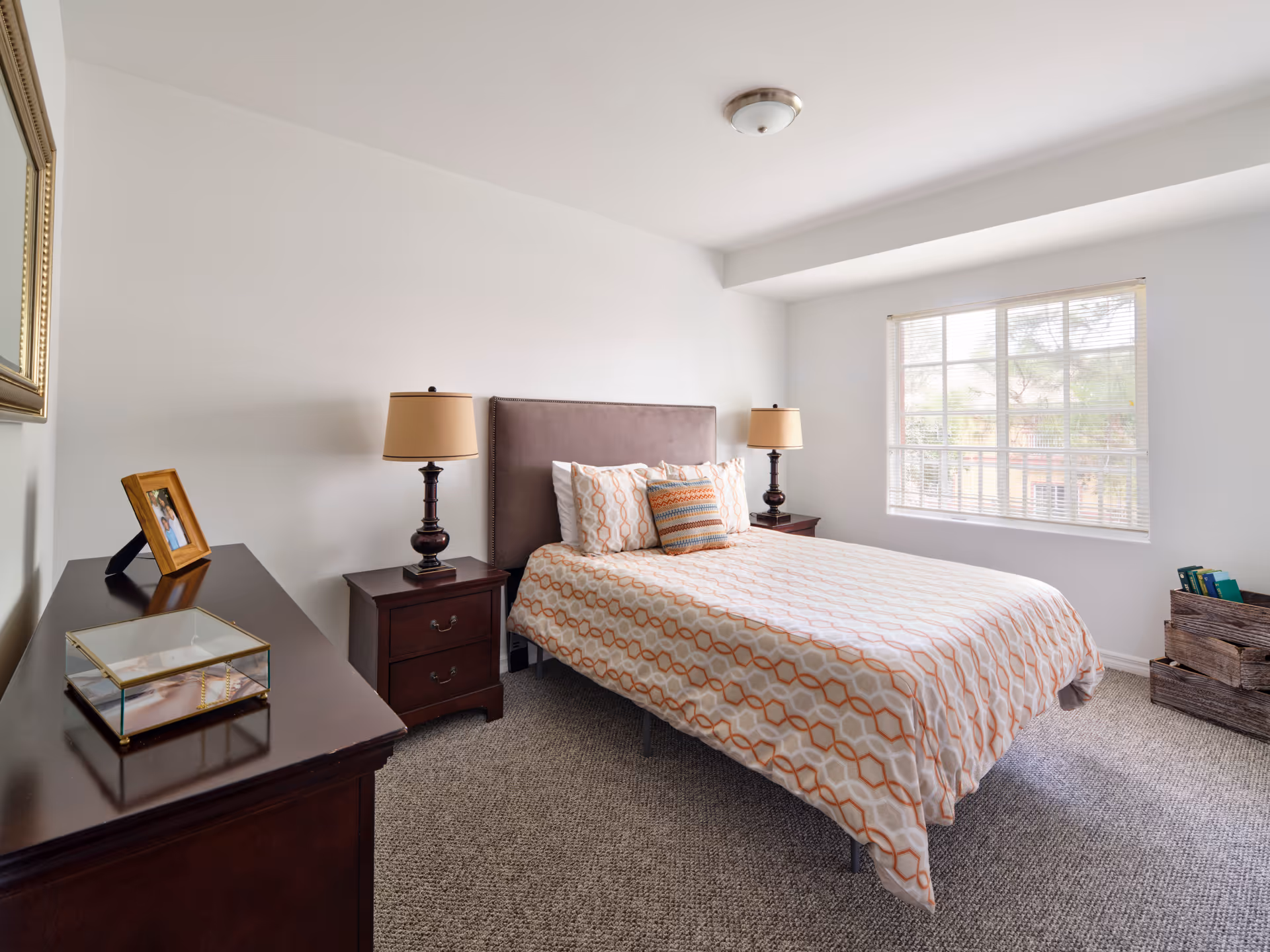 A bright bedroom with a large window letting in natural light. The room features a bed with a patterned bedspread and multiple pillows, two dark wooden nightstands each with a lamp, a dark wooden dresser with a framed photo and a glass box on top, and a woven basket with books near the window. The walls are white and the floor is carpeted.