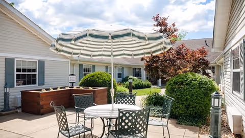 Outdoor patio area with a round table and four metal chairs under a large green and white striped umbrella. Surrounding the patio are bushes, a tree with red leaves, and white buildings with green shutters under a partly cloudy sky.