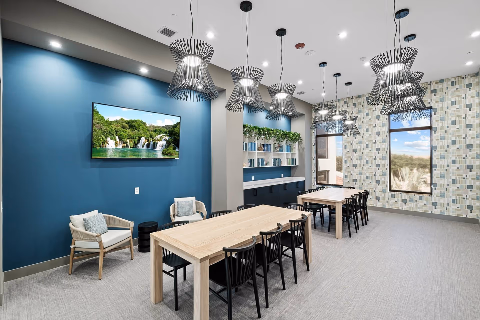 A modern dining area with two light wooden tables surrounded by black chairs. The room features a blue accent wall with a mounted flat-screen TV displaying a waterfall scene, two cushioned armchairs, and a built-in shelf with books and greenery. Large windows with a mosaic tile wall allow natural light to enter. Several contemporary pendant lights hang from the ceiling.