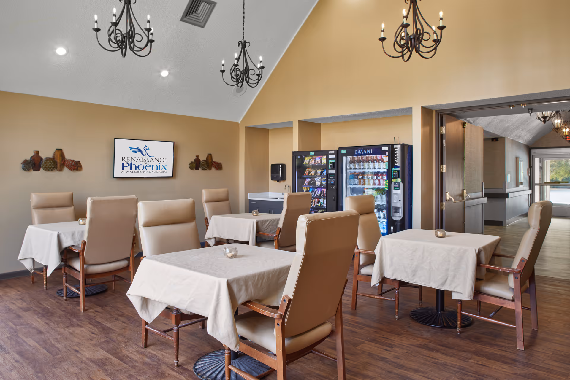 Dining room with tables draped in beige cloths, cushioned chairs, chandeliers, and a vending machine against the far wall.