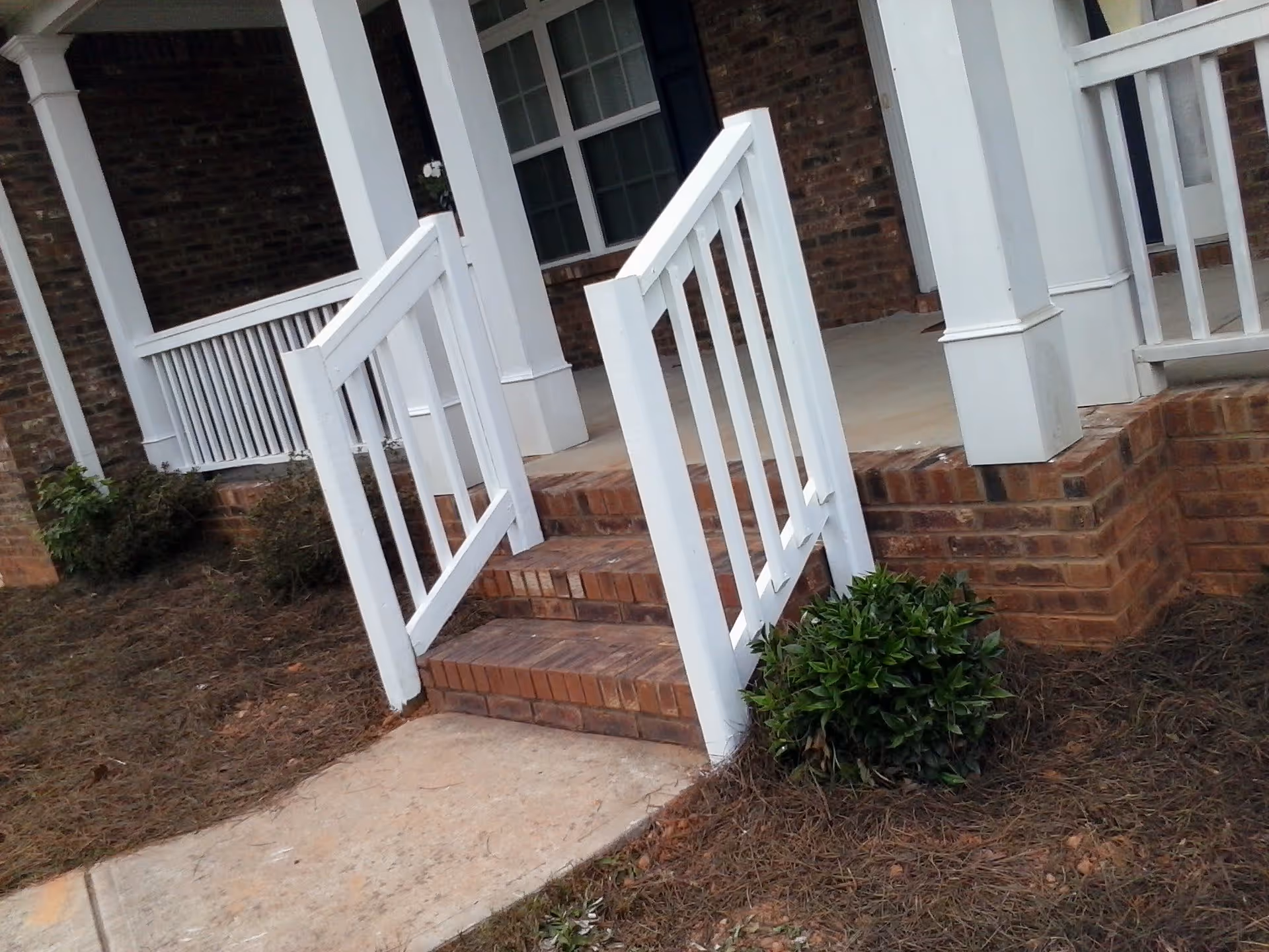 A small brick porch with three steps leading up to it, featuring white wooden railings and columns. There is a small green shrub at the base of the steps and a concrete pathway leading to the porch. The building exterior is made of red brick.