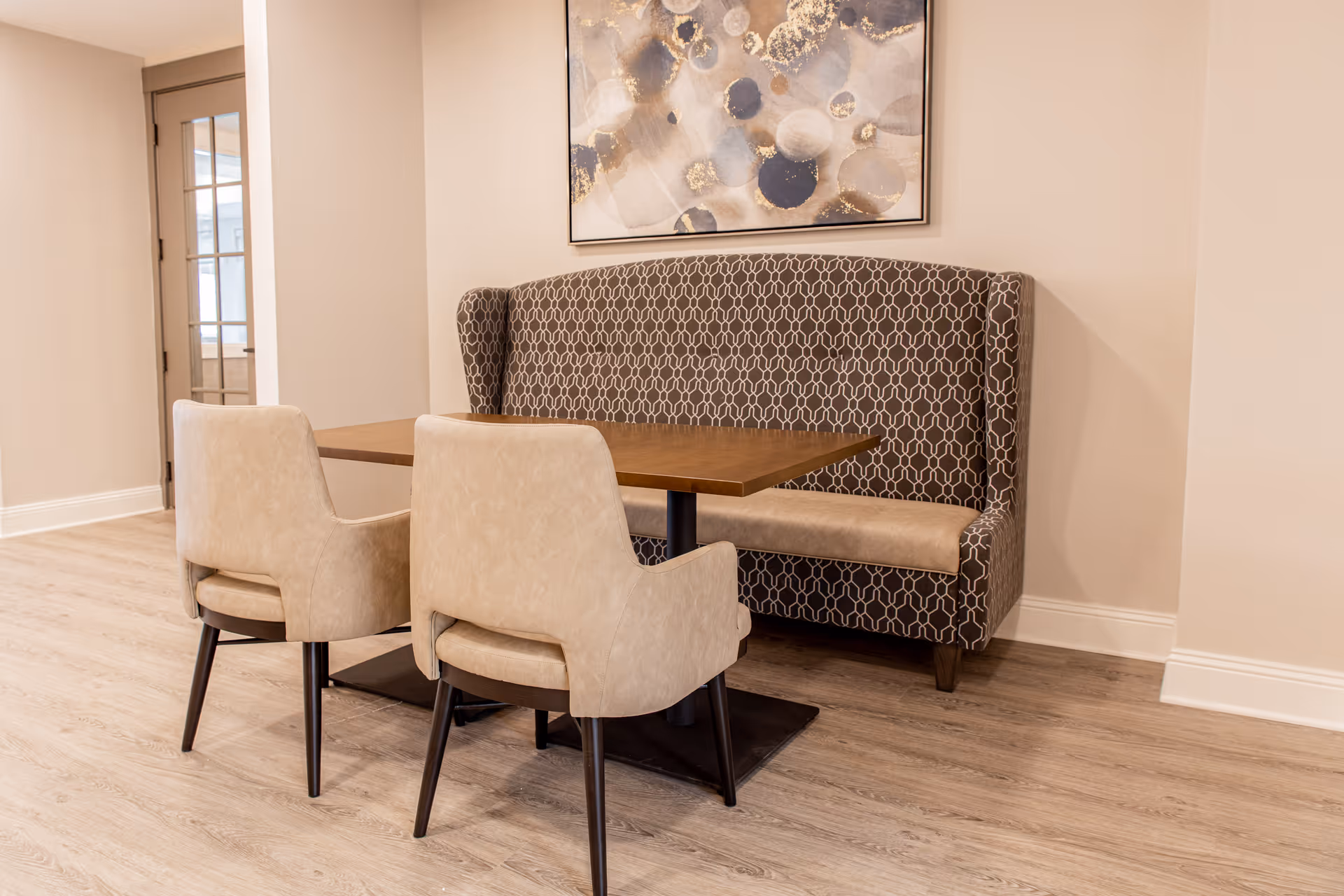 A dining nook with a patterned upholstered banquette, two beige chairs, and a wooden table beneath abstract wall art.