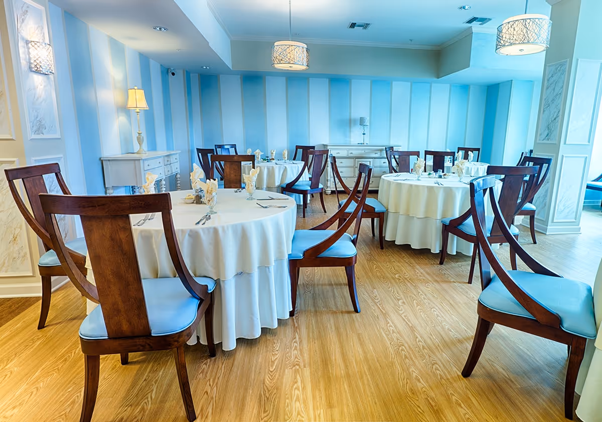 A bright dining room with round tables covered in white tablecloths, each set with napkins, glasses, and silverware. The room features wooden chairs with blue cushions, light blue striped walls, wooden flooring, and soft lighting from ceiling fixtures and a table lamp.