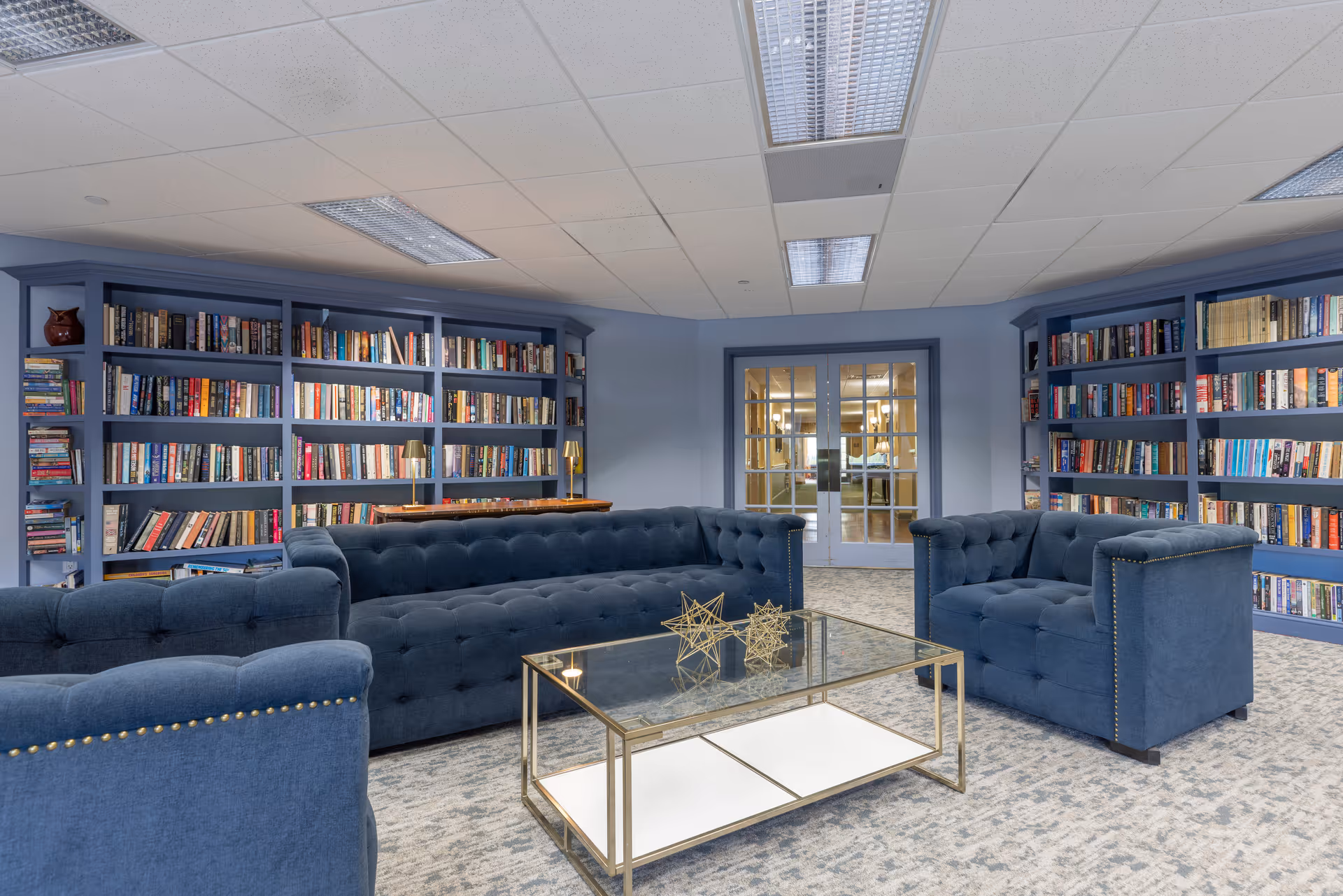 A cozy library room with blue bookshelves filled with books along the walls. In the center, there are blue upholstered sofas and armchairs arranged around a glass coffee table with decorative gold geometric sculptures. The room has a carpeted floor and a ceiling with fluorescent lights. Double glass doors are visible at the back of the room.