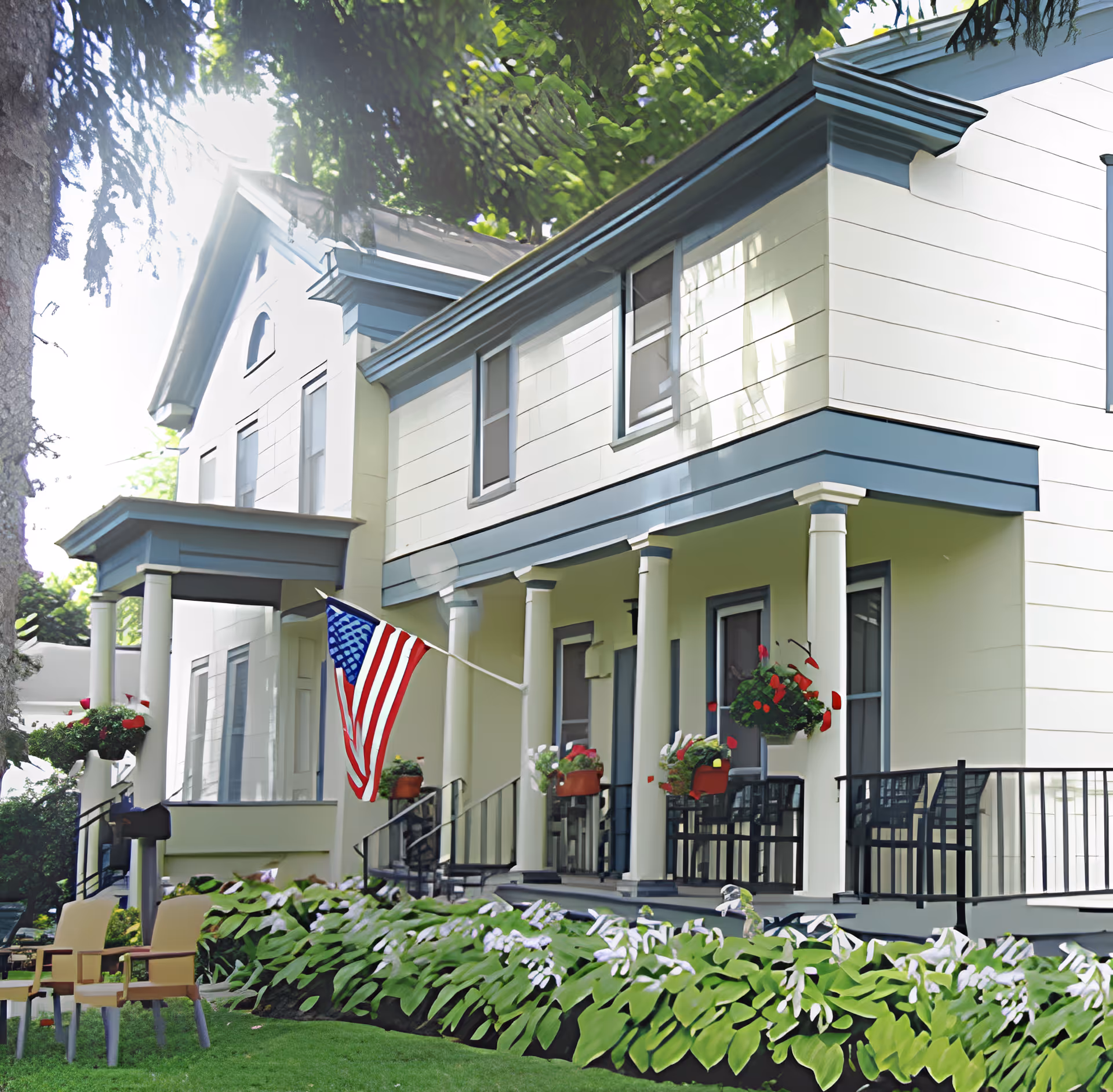 Exterior view of a two-story assisted living facility with white siding and blue trim. The building features a covered porch with white columns, hanging flower baskets, and an American flag mounted near the entrance. There are green plants and flowers along the front garden bed and two chairs on the lawn.