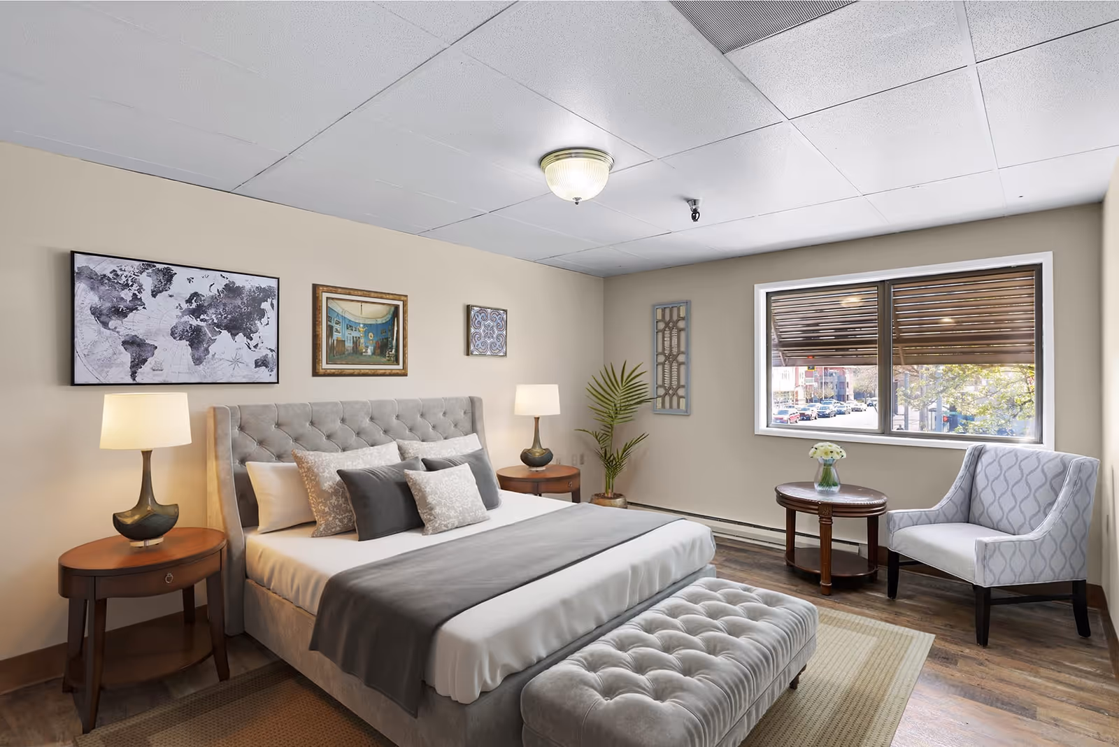 A well-lit bedroom with a large bed featuring a tufted headboard and multiple pillows. There are two wooden nightstands with lamps on either side of the bed. On the wall above the bed are three framed pictures, including a world map. A cushioned bench is at the foot of the bed. To the right, there is a window with a view of a street outside, a small round wooden table with a vase of flowers, and a patterned armchair. The floor is wooden with a beige rug partially visible.