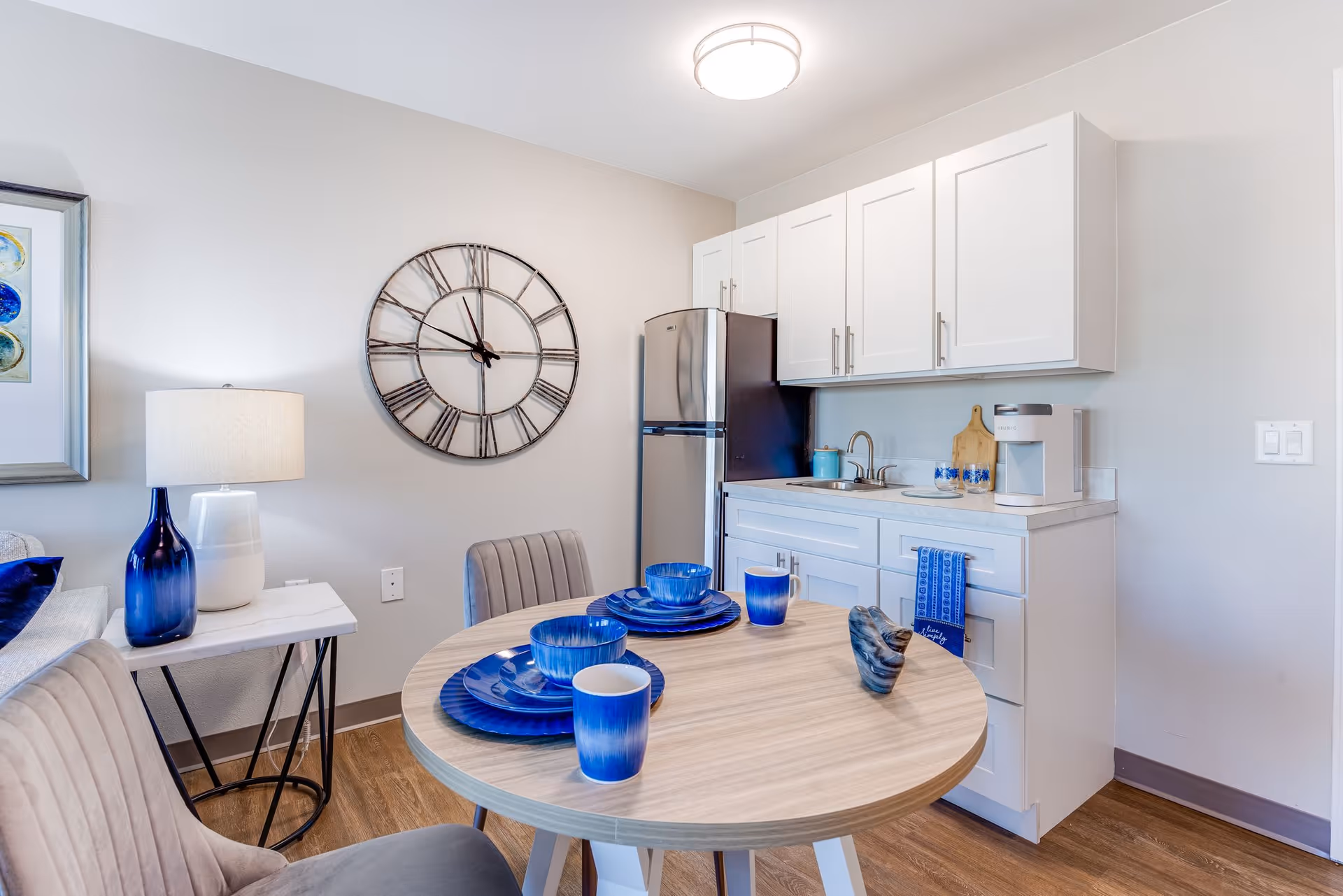 A small kitchen and dining area featuring white cabinets, a stainless steel refrigerator, a round wooden table set with blue dishes and cups, a gray upholstered chair, a side table with a blue vase and white lamp, and a large wall clock with Roman numerals.