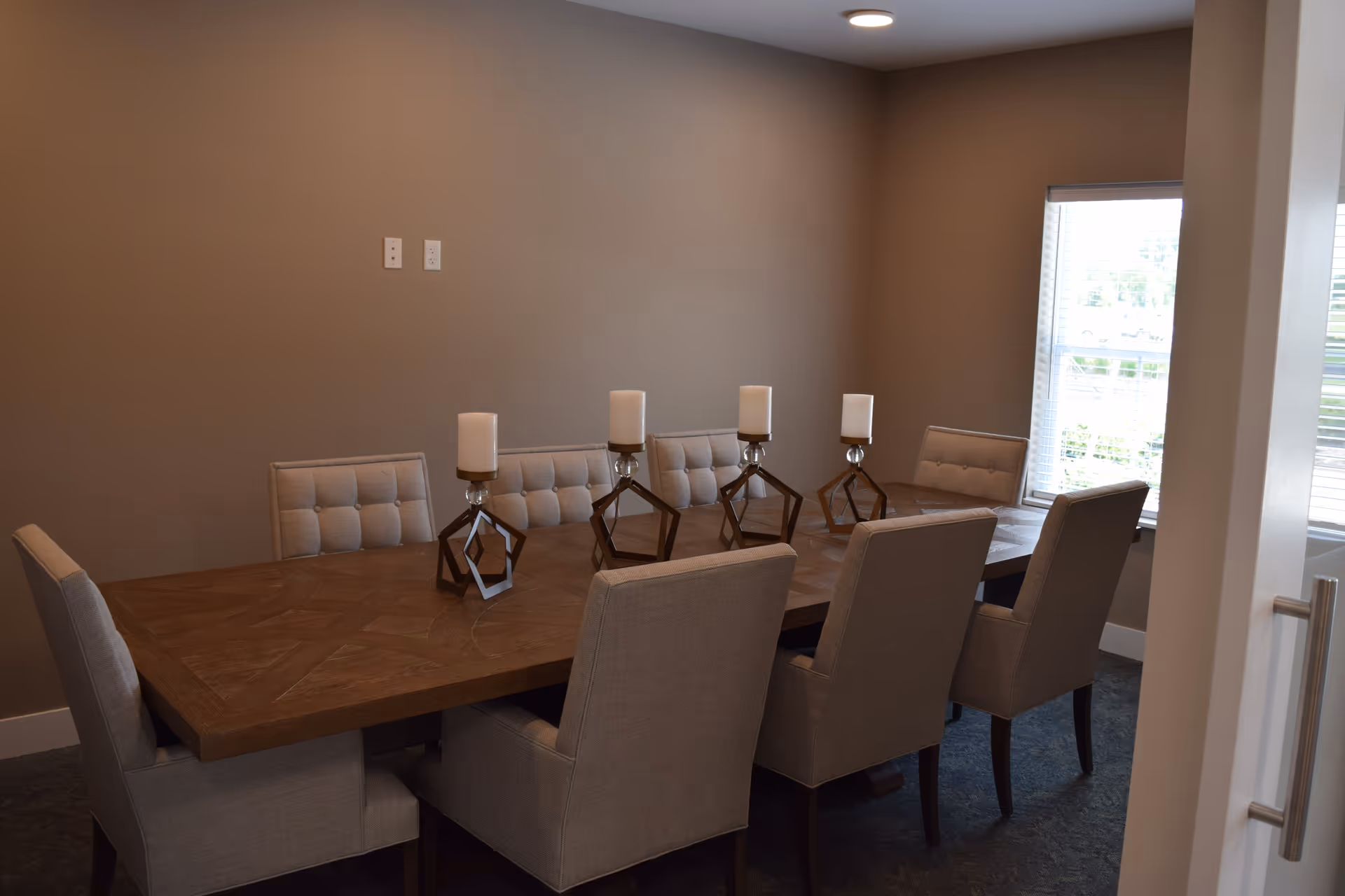 A dining room with a large wooden table surrounded by upholstered chairs and four geometric candle holders, with a window letting in natural light.