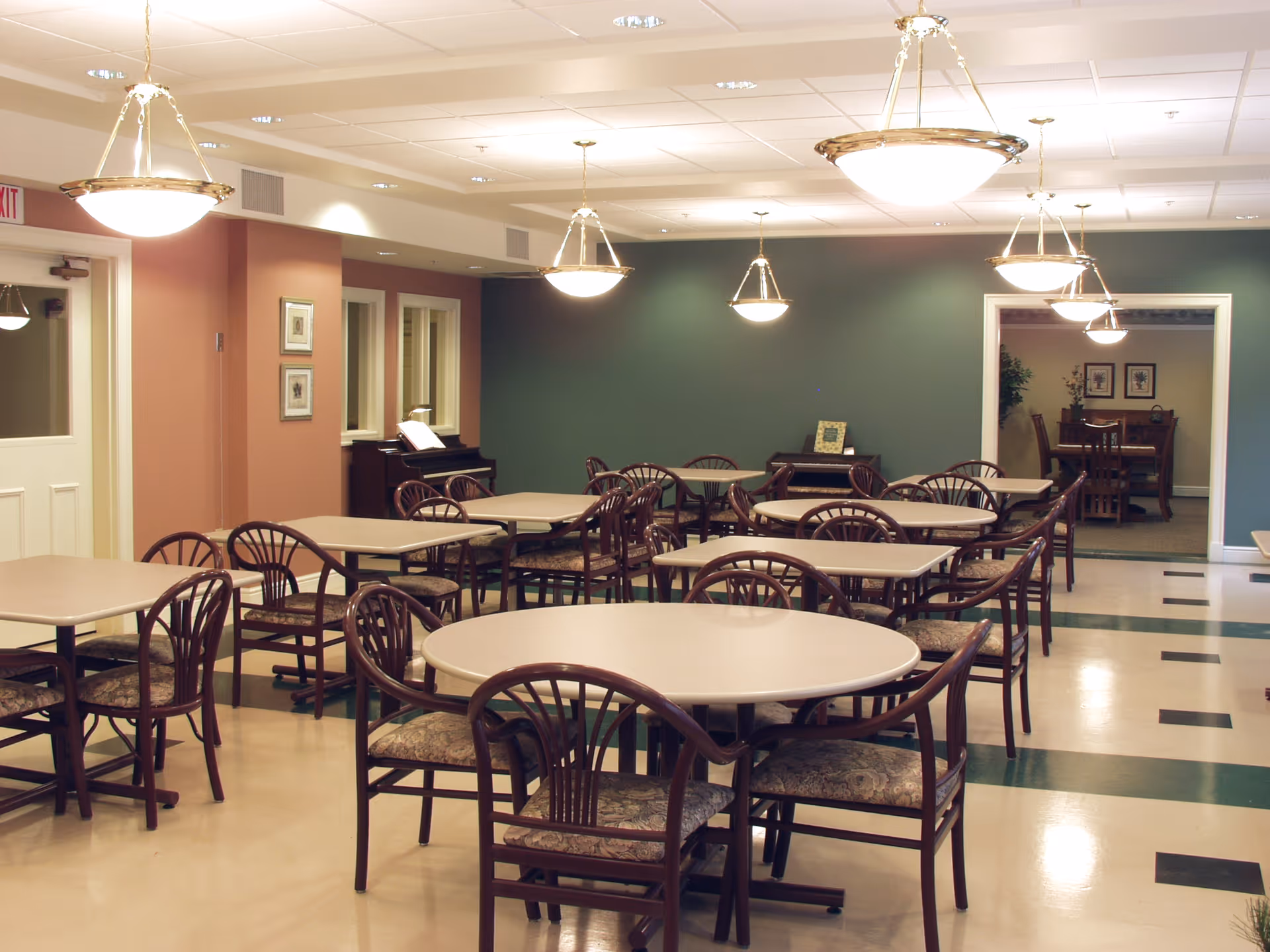 A dining room in The Oaks Retirement Residence with multiple tables and chairs arranged neatly. The room has soft lighting from hanging ceiling lamps, a green accent wall, and a piano in the corner. There is an adjoining room visible through a doorway with additional seating and framed artwork on the walls.