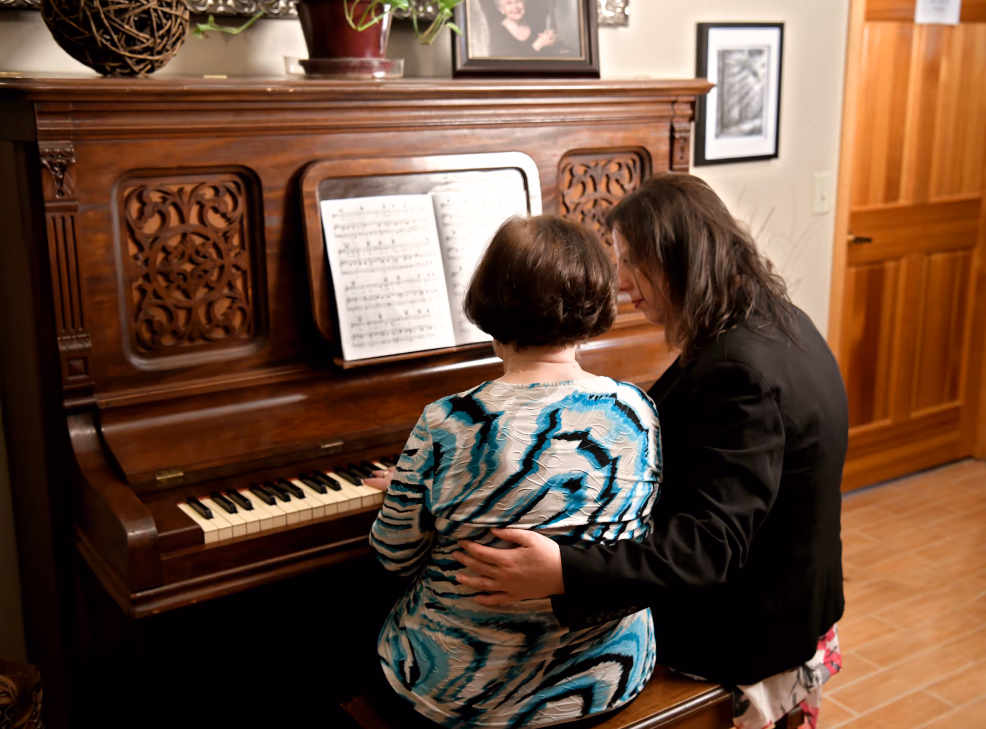 Two women sitting at an ornate wooden piano with sheet music displayed. One woman is playing the piano while the other, seated beside her, has her arm around her in a supportive gesture. The room has warm lighting, a wooden door, framed pictures on the wall, and decorative items on top of the piano.