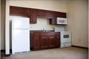 Kitchenette with a white refrigerator, stove, microwave and dark wood cabinets above and below a sink in an otherwise empty room.