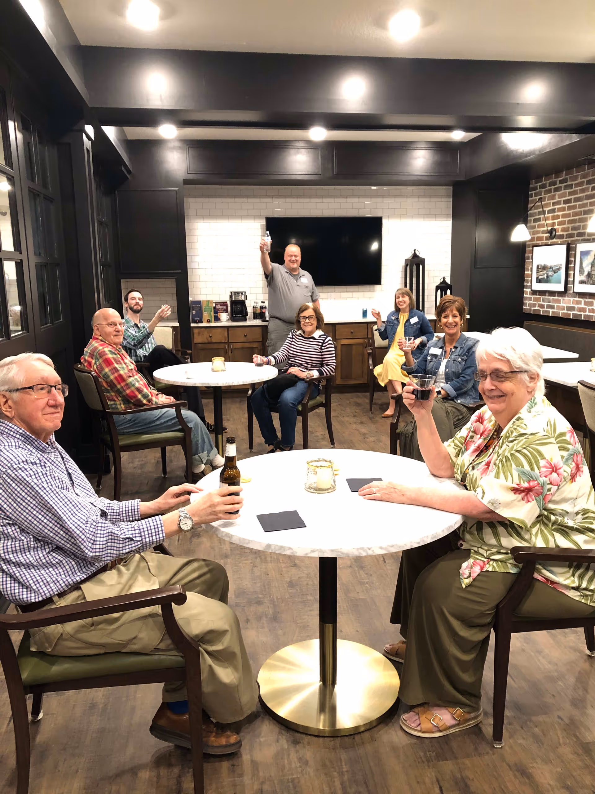 A group of seven adults, mostly seniors, sitting and standing around tables in a well-lit room with dark wood accents and a large TV mounted on a white tiled wall. They are smiling and raising drinks in a toast, creating a warm and social atmosphere.