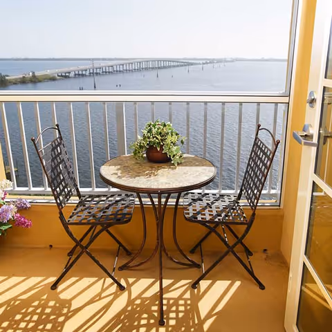 A small balcony with a round table and two metal chairs overlooking a large body of water and a distant bridge. There is a potted plant on the table and flowers on the left side of the balcony. The balcony has a railing and a door leading inside.