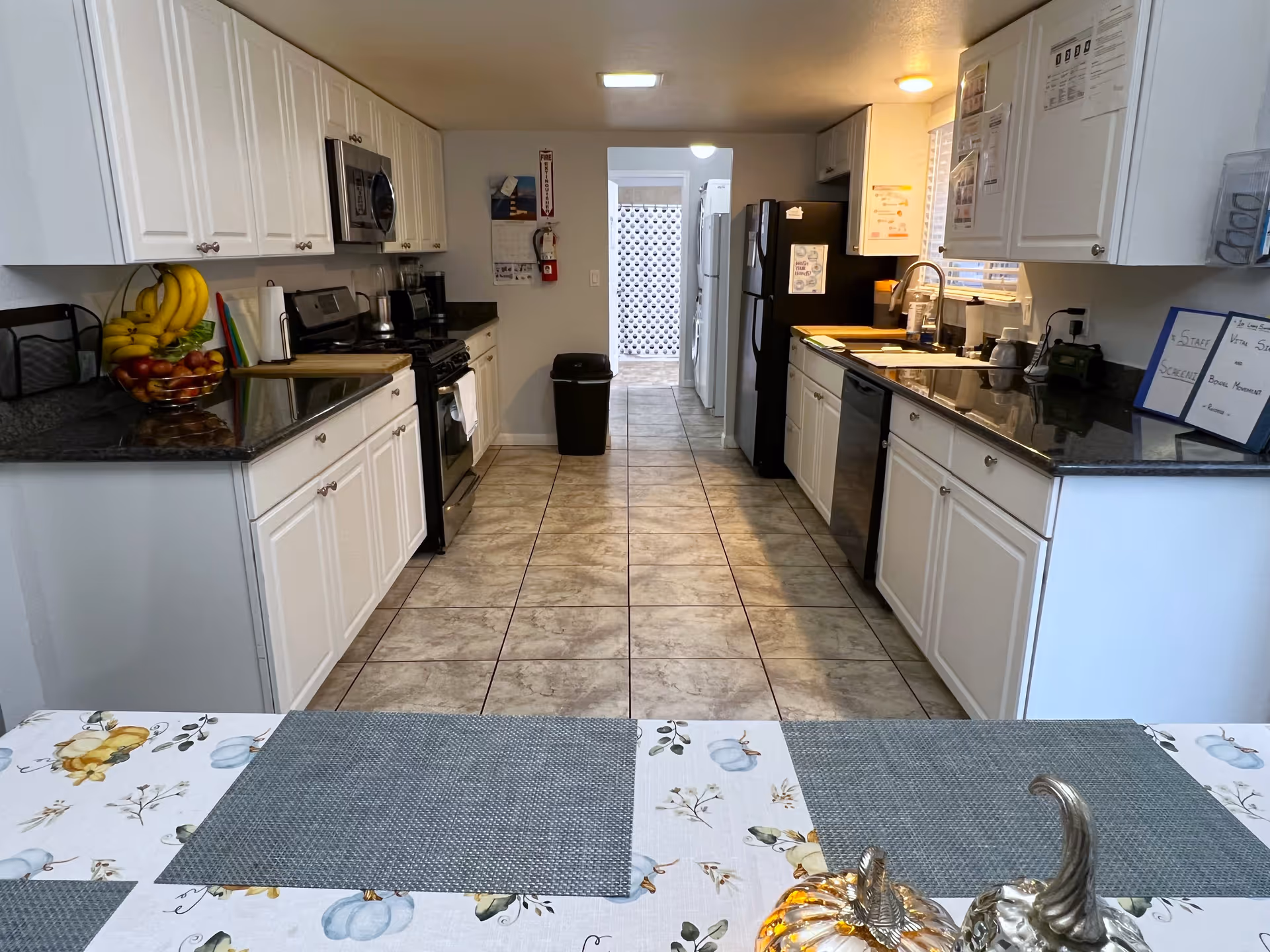 A clean, well-lit kitchen with white cabinets and black countertops on both sides. The kitchen features a stove, microwave, dishwasher, and two refrigerators. There is a tiled floor and a table with a floral tablecloth and placemats in the foreground. Various kitchen items like a fruit bowl with bananas and apples, a fire extinguisher, and a few notes or signs are visible.