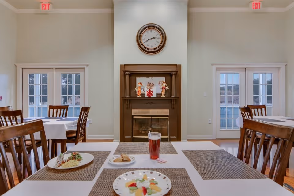 Dining room with wooden chairs and tables covered with white tablecloths and brown placemats. Plates with food and a glass of beverage are on the table in the foreground. A fireplace with decorative items and a round clock above it is centered on the far wall. Two sets of French doors with glass panes are on either side of the fireplace, and exit signs are visible above each door.