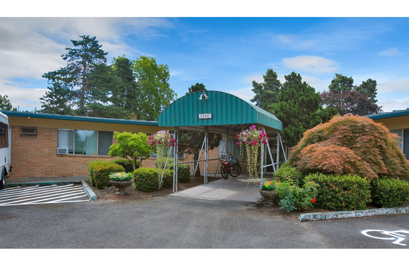 Front entrance of a single-story brick building with a green arched canopy, hanging flower baskets, and landscaped shrubs.