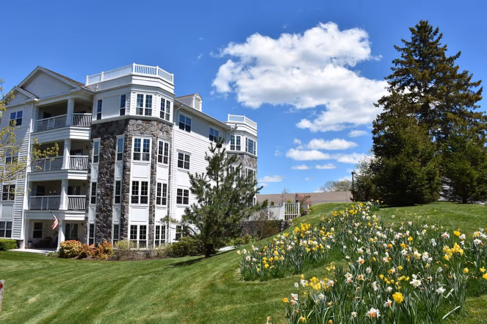 Exterior view of a multi-story senior living facility building with white siding and stone accents, surrounded by a well-maintained lawn and flower beds with yellow and white flowers under a blue sky with scattered clouds.