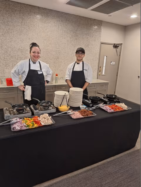 Two chefs standing behind a table set up with cooking stations, plates, and trays of chopped vegetables and meats in a kitchen or food preparation area.