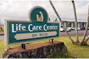 Green sign reading 'Life Care Center of Hilo' on a stone base in front of the facility, with palm trees and the building visible behind it.
