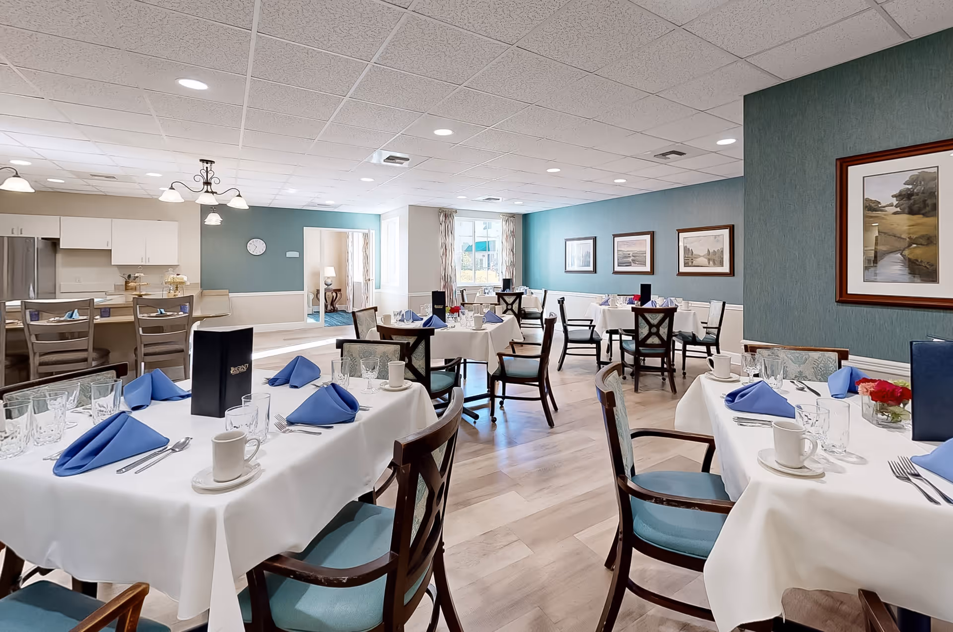 A bright and elegant dining room with several round tables covered in white tablecloths, each set with blue folded napkins, glassware, cups, and silverware. The room features wooden chairs with blue cushions, teal walls adorned with framed landscape paintings, and a kitchen area with a refrigerator and cabinets in the background. The space is well-lit with ceiling lights and natural light from windows.